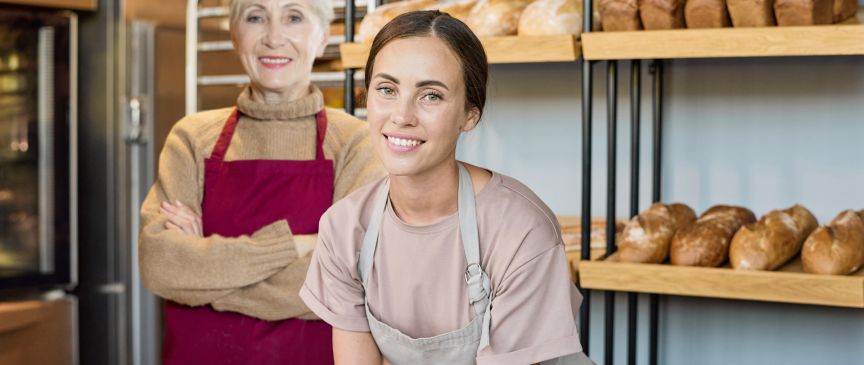 Zwei Frauen in roten und beigen Schürzen stehen in einer Bäckerei, im Hintergrund Brotlaibe auf Holzregalen.