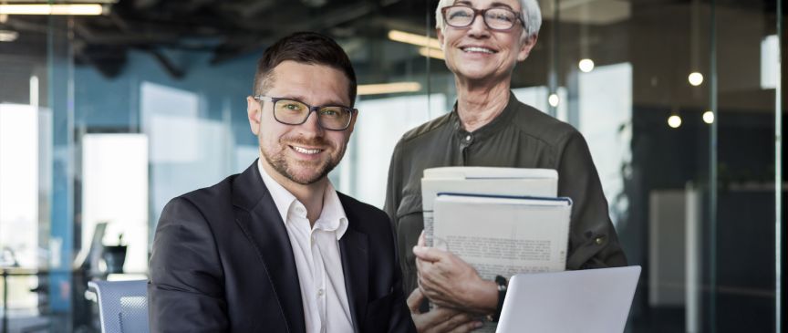 Ältere Unternehmerin steht hinter einem sitzenden jüngeren Mann an einem Notebook