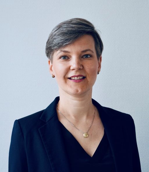 Frau mit kurzem grau-braunem Haar, dunklem Blazer und Goldkette lächelt vor hellem Hintergrund.