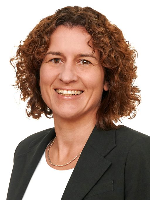 Frau mit lockigem, rotbraunem Haar, dunklem Blazer und Halskette lächelt vor weißem Hintergrund.