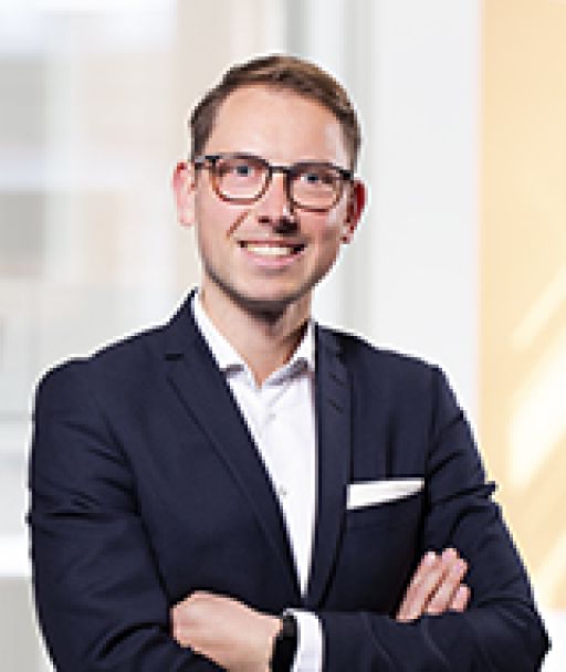 Mann mit Brille im dunklen Blazer, Arme verschränkt, vor hellem Hintergrund, professionelles Porträtfoto.