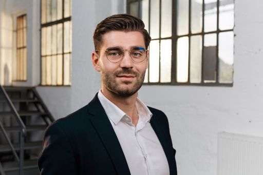 Mann mit Brille und Bart, dunklem Sakko und weißem Hemd, steht vor einem Industriegebäude mit Fenstern.