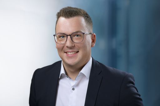 Mann mit Brille und kurzen Haaren, trägt weißes Hemd und dunkles Sakko, lächelt vor hellem Hintergrund.