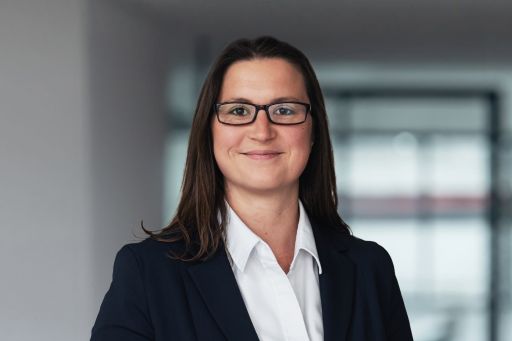Frau mit langen braunen Haaren und Brille in weißer Bluse und dunklem Blazer lächelt in die Kamera.