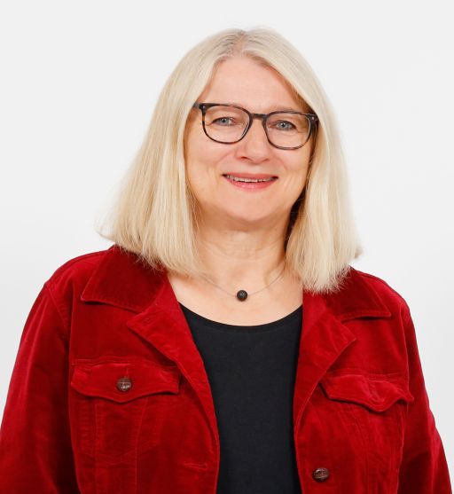 Lächelnde Frau mit blonden Haaren und Brille, roter Jeansjacke und schwarzem Shirt, vor weißem Hintergrund.