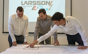 Drei Männer beugen sich über großformatige technische Zeichnungen auf einem Tisch vor einer Leinwand mit Logo.