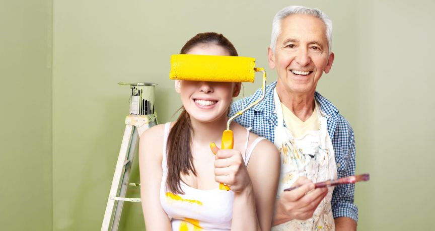 erfolgreiche Unternehmensübergaben sichern Unternehmen und Arbeitsplätze älterer Malermeister steht hinter einen jungen Frau, die sich eine Farbrolle vor die Augen hält.